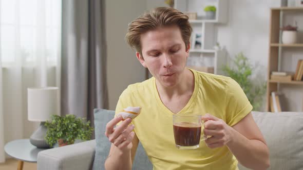 Cheerful Guy Eats a Donut at Home in the Living Room and Drinks Coffee