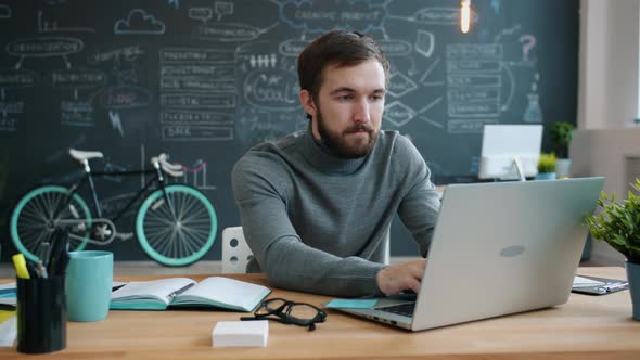 Serious Guy Using Laptop Typing Sitting at Table in Creative Cozy Office Concentrated on Job