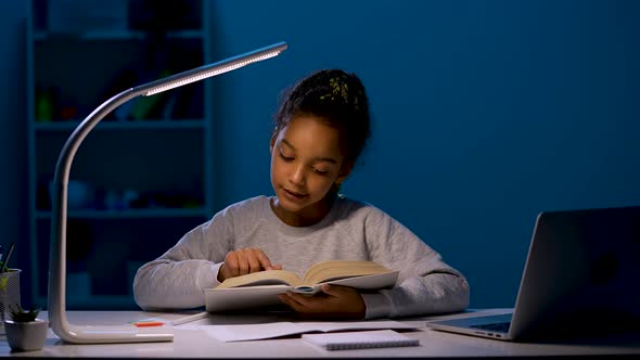 An African American Schoolgirl Under the Light of a Night Lamp Does Her Homework Reads a Book Aloud