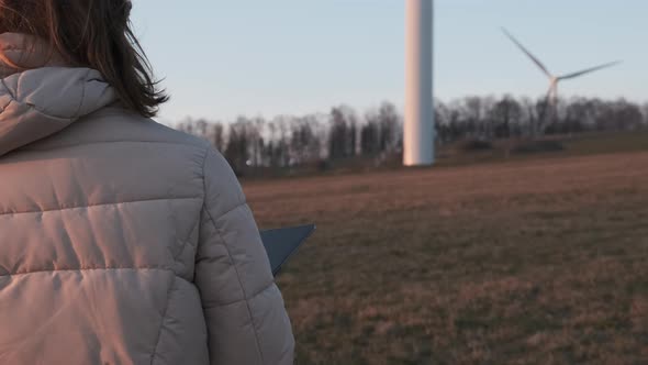 Female Engineer in Orange Vesta Goes to Wind Turbines with a Tablet to Checks Their Operation