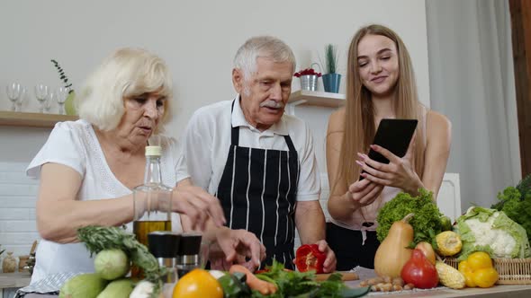 Mature Grandparents Couple Cutting Vegetables for Salad, Listening Recipe From Girl with Tablet