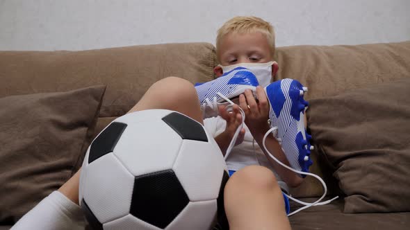 A Small Sad Boy in a Medical Mask Sits on a Sofa with a Soccer Ball and Cleats