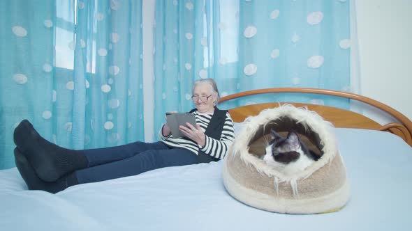 Old Woman Holding Tablet PC Lies on Bed Near Spoted Kitten