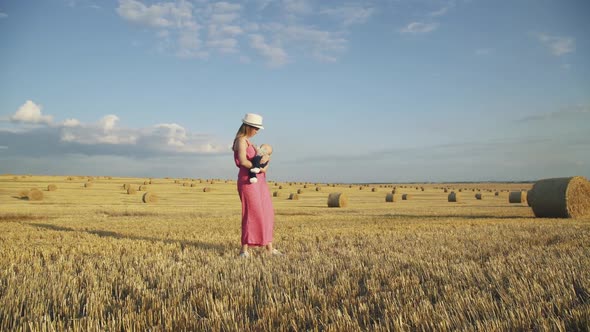 Stylish Mother Lulling Baby Son in Hands in Hay Field During Hot Summer Day