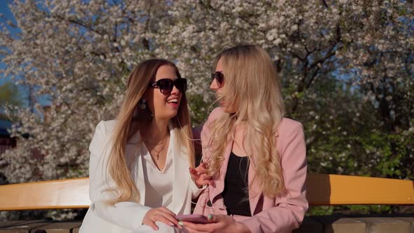 Two females persons looking at smartphone smile and laugh outside. Young adult women in sunglasses