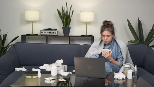 Sick Young Woman Sitting on Sofa with Cup of Tea and Laptop