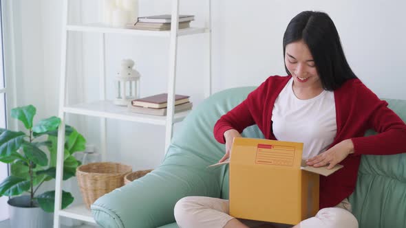 Excited young asian girl consumer holding opening cardboard box sit on sofa at home	