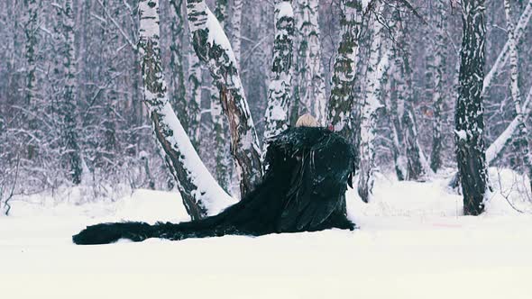 Woman in Phoenix Suit with Wings Squats at Tree in Forest