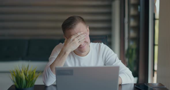 Upset Young Man Covering Face with Palm in Despair After Reading Bad News on Laptop