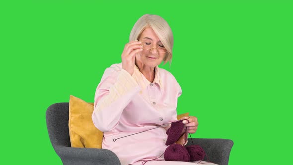 Elderly Woman in Glasses Knits Clothes and Reacts on Something Behind the Camera on a Green Screen
