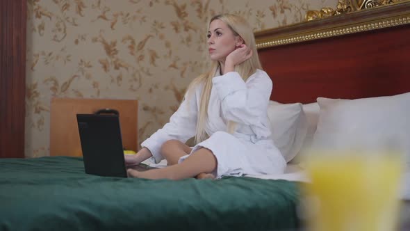 Young Gorgeous Slim Caucasian Blond Woman Sitting on Bed in Luxurious Hotel Room Typing on Laptop