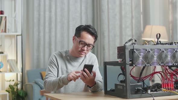 Asian Man Smiling And Shaking His Head While Using Smartphone With Mining Rig Mining Cryptocurrency