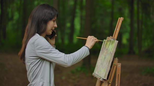 Middle Side View of Girl Artist Painting a Landscape in the Park and Talking By the Phone