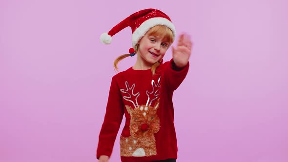 Child Girl in Christmas Sweater Waves Hand in Hello Gesture Welcomes Someone to Celebrate New Year