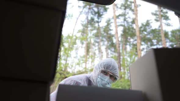 A Volunteer in a Protective Suit Loads Boxes Into the Trunk of a Car.