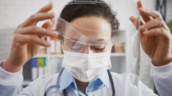 Doctor Woman in Protective Mask is Putting on a Face Shield and Looking at you While Standing in