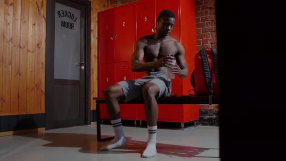 Africanamerican Athletic Young Man Sitting in the Locker Room and Drinking Water From the Plastic