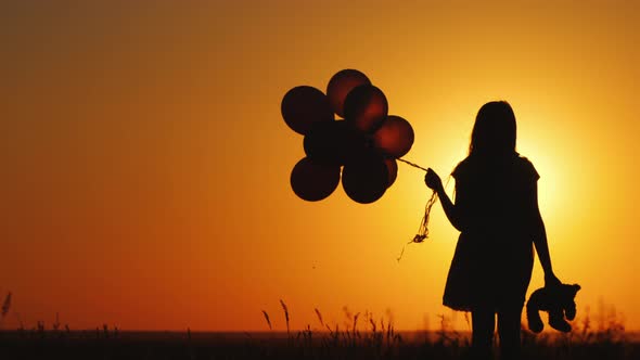 A Child with Balloons and a Teddy Bear in His Hand is Standing at Sunset