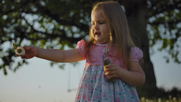 A Little Girl in a Dress Invites Her Little Sister To Blow Soap Bubbles.