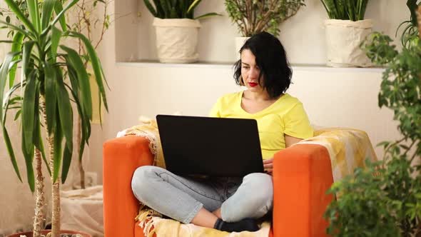 Woman using, search a laptop on a orange armchair at home