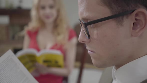 Young Concentrated Man in Glasses Reading the Book in the Foreground While Pretty Blond