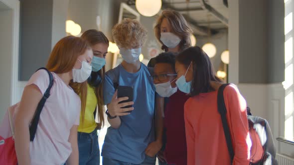 Group of Classmates Wearing Protection Medical Mask Looking at Mobile Phone Standing in Corridor