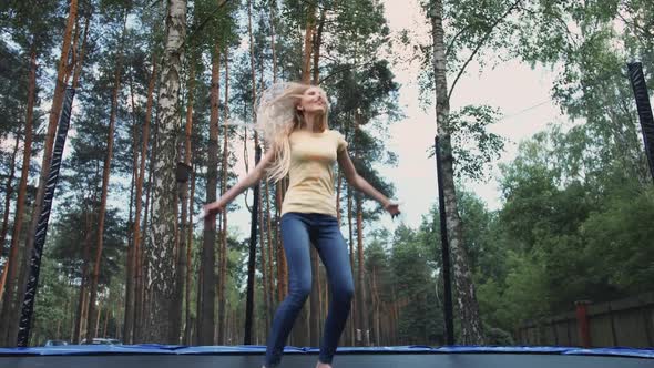 Happy Female Jumping on Trampoline. Beautiful Young Blond Barefoot Woman in Light Summer Shirt and