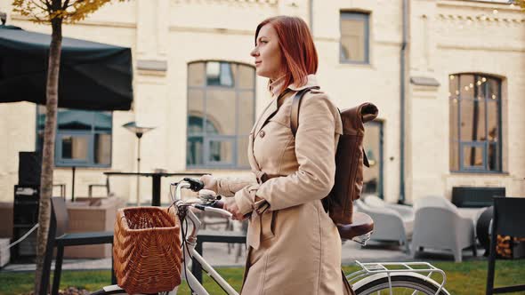 Young Redhead Female is Riding Cruiser Bike By Deserted City Square Decorated with Lights