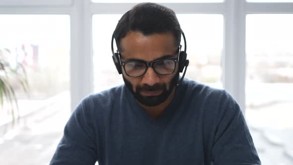 Front View of Midage Indian Male in Casual Wear Using Headset and Modern Laptop