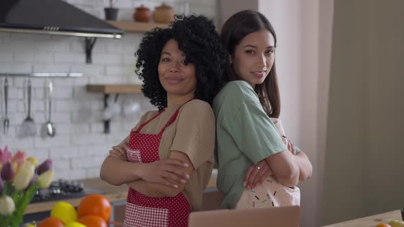 Middle Shot of Confident Young Women in Aprons Standing Back to Back Crossing Hands Smiling Looking