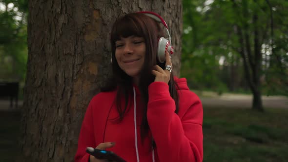 Woman, standing near a tree in the park, uses a smartphone and listens to music with headphones.