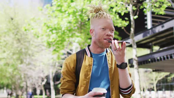 Happy albino african american man with dreadlocks in park drinking coffee talking on smartphone