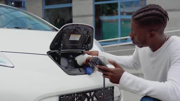 A Young African American Man Connects an Electric Car to the Charger and Adjusts the Process of