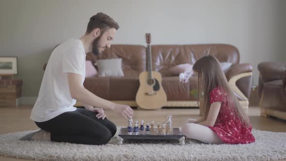 Young Bearded Father and Little Girl with Long Hair Playing Chess Sitting on the Floor on Fluffy