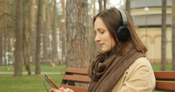 Attractive woman puts on headphones and turns on music while sitting on bench in autumn park 