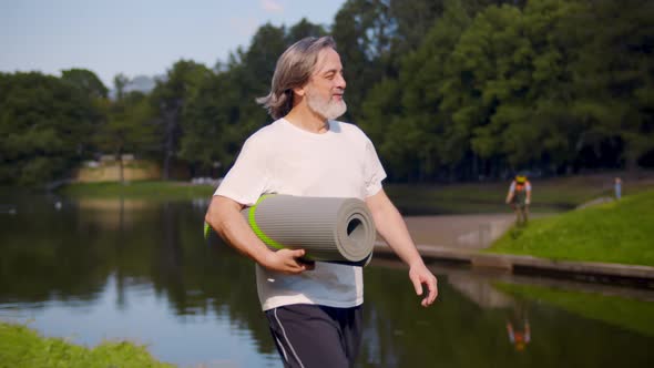 Smiling Mature Man Holding Yoga Mat and Walking in Summer Park