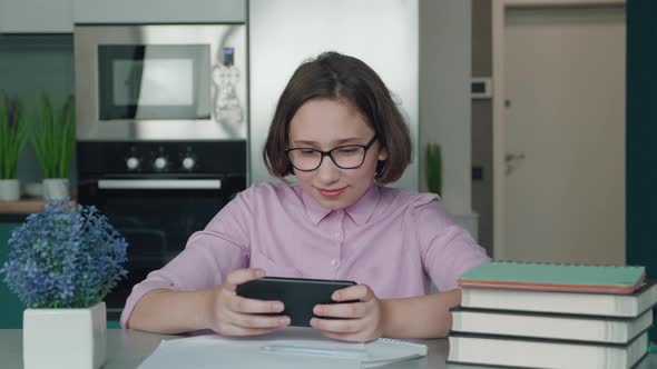 Little Girl with Glasses Sitting Into the Desk Using Smartphone Watching Video Child is Bored