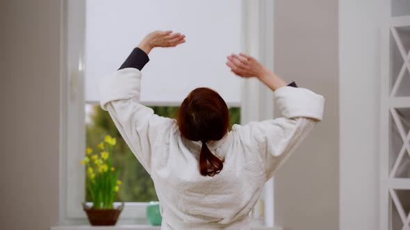 Back View Slim Mature Caucasian Woman in White Bathrobe Stretching in the Morning at Home Indoors