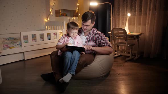 Happy Laughing Boy with Young Father Sitting in Bedroom at Night and Playing Games on Tablet