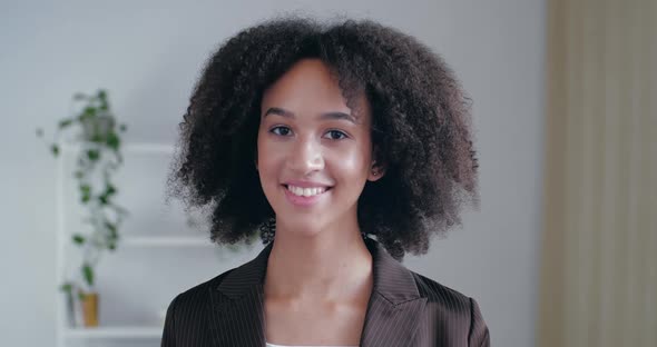 Сurly-haired Young American Woman Stands in Office at Home in Room in Front of Camera, Smiles