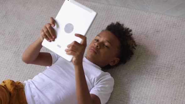 African American Boy is Using Electronic Device While Lying on Floor in Apartment Room Spbi