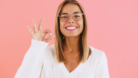 Young Lady in Glasses and White Jumper is Showing Hand Sign Okay Smiling While Posing on Pink