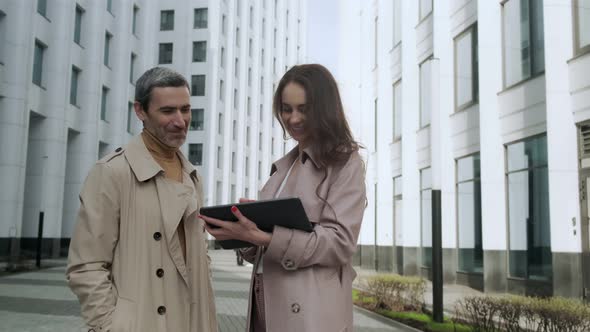 A beautiful girl with a tablet approaches the head of a man standing near office buildings