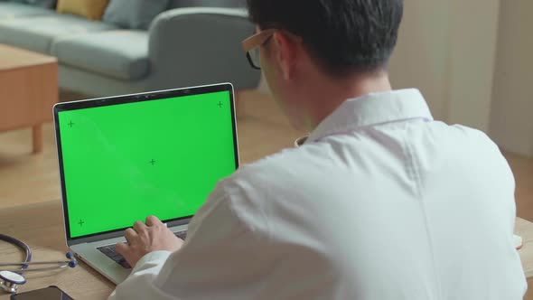 Male Doctor Typing On Laptop Computer With Green Screen Display At Home Office