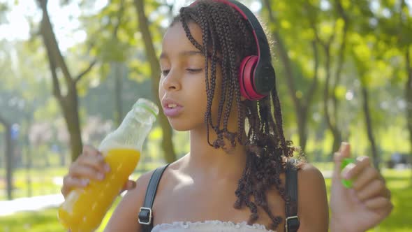 Close-up of Positive Little Girl in Headphones Drinking Orange Juice in Sunny Park. Portrait of