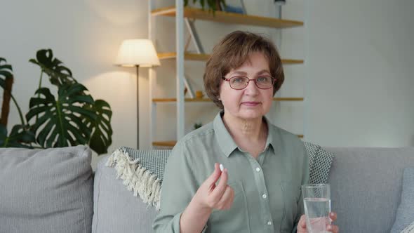 Beautiful Senior Woman with a Pill and a Glass of Water in Her Hands Sits on a Sofa at Home