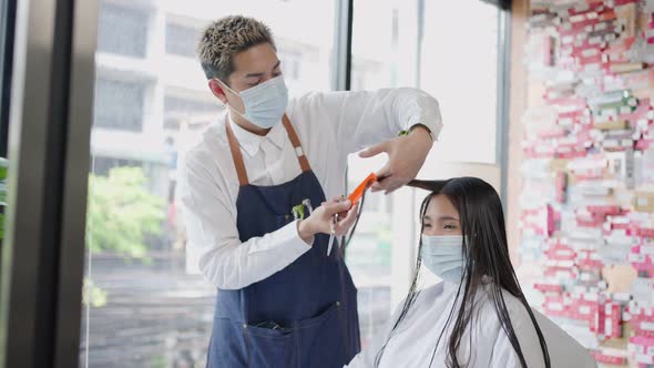 Asian male hairstylist wear mask, combing and cutting young woman customer's hair in barber salon.