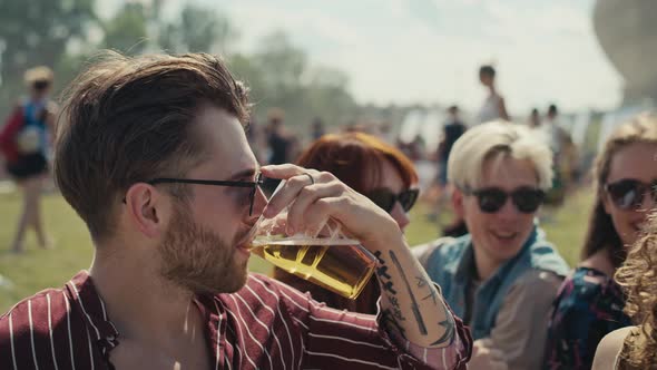 Group of friends sitting on grass together at music festival and drinking beer while chatting. Shot