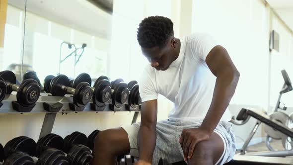 Young African American Man Sitting and Lifting a Dumbbell Close to the Rack at Gym