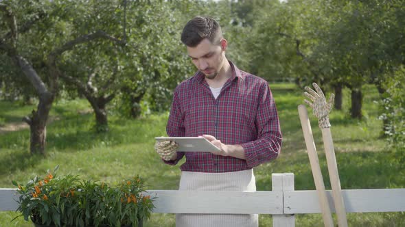 Young Farmer in Garden Gloves Using the Tablet Standing Behind the Fence in the Foreground